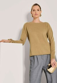 Frau in beigem Pullover und grauen Plisseehosen lehnt an einem weißen Podest, hält eine goldene Clutch und trägt große goldene Ohrringe und Armbänder.