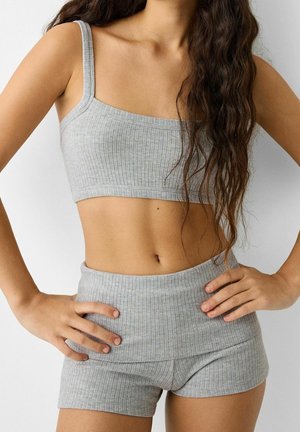 Frau trägt ein graues, geripptes Crop-Top und passende hoch taillierte Shorts, steht mit den Händen in der Hüfte vor weißem Hintergrund.