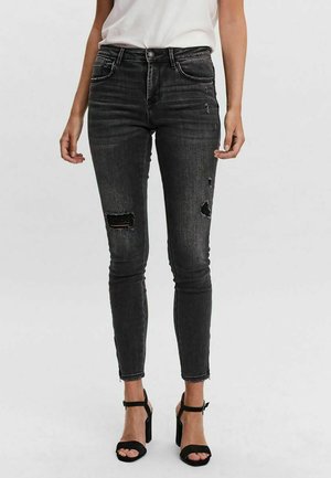 Kobieta ubrana w czarne rozdarcia skinny jeans, biały top i czarne sandały na wysokim obcasie stoi na jednolitym tle.