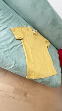 Rumena polo majica iz gladkega materiala, z kratkimi rokavi in ovratnikom, leži raztegnjena na teksturirani svetlo modri kavč.