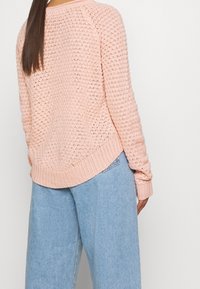 Vaaleanpunainen neulepaita, jossa on teksturoitu kuvio, ribattu hihansuu ja helma, sekä rento malli, yhdistettynä vaaleansinisiin leveälahkeisiin farkkuihin.