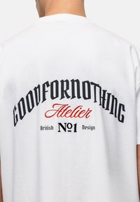Witte katoenen T-shirt met zwarte en rode tekst: "GOODFORNOTHING Atelier British No1 Design" in vette, gestileerde lettertypen op de achterkant.