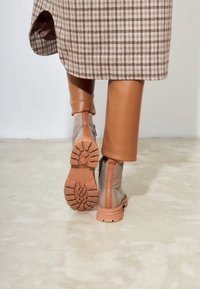 Manteau à carreaux marron sur un pantalon en cuir ajusté marron, mettant en valeur des bottines beiges avec des semelles texturées et des accents marron contrastants.
