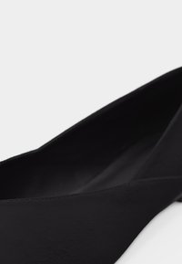 Chaussure plate pour femme noire, à bout pointu et matière supérieure lisse, présentée en angle sur un fond blanc.