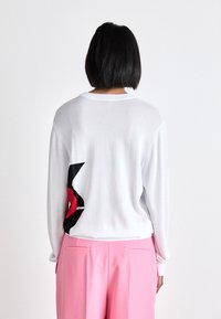 Personne aux cheveux courts noirs portant un pull blanc avec un motif noir et rouge dans le dos et un pantalon taille haute rose, vue de dos.