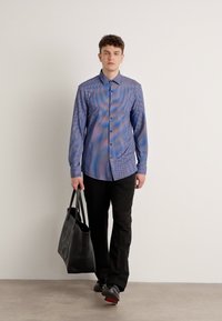 Camisa de botones azul con estampado, pantalones negros, llevando una bolsa tote negra. Tejido suave con un diseño geométrico y mangas largas.