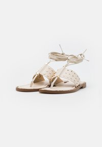 Sandali piatti beige con accenti di borchie, design open toe e lacci alla caviglia. Realizzati in materiale morbido con suola piatta e dettagli in hardware dorato.