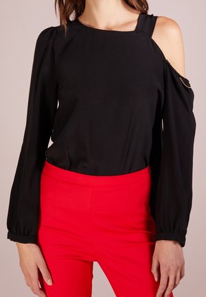 Vrouw draagt een zwarte cold shoulder blouse met lange mouwen en felrode hoge taille broek, staat met ontspannen armen langs het lichaam.