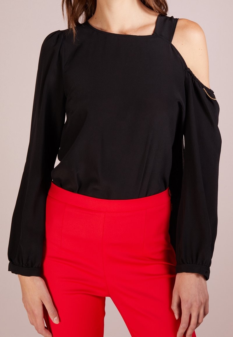 Femme portant un chemisier noir à épaules dénudées avec manches longues et un pantalon taille haute rouge vif, debout, les bras détendus le long du corps.