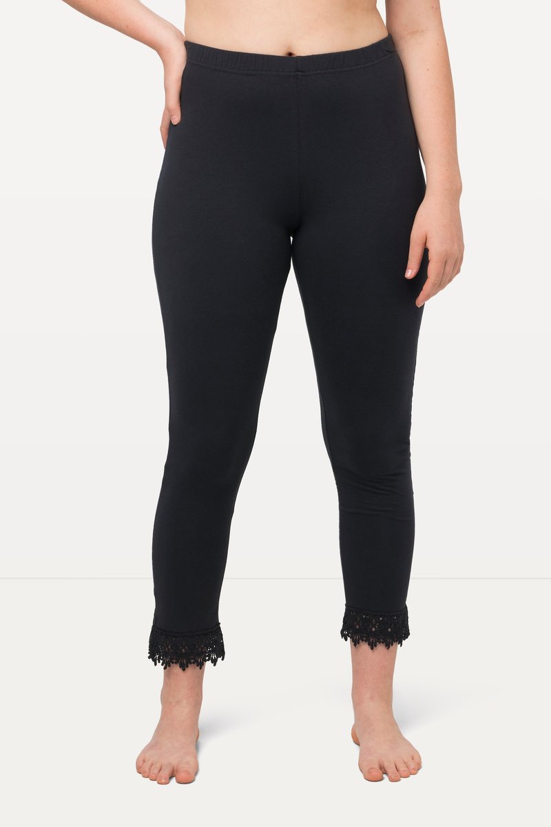Kvinde iført sort tætsiddende leggings med blondekant ved anklen, stående barfodet mod en neutral baggrund.
