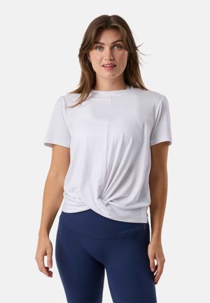 Woman wearing a white short-sleeve twist-front top and navy blue fitted pants, standing against a plain light background.