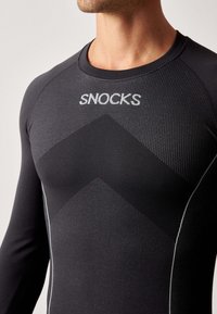 Czarna długosleeve koszulka sportowa z teksturowanej tkaniny, z szarym wzorem w zygzak i logo "SNOCKS" w metalicznej srebrnej barwie wzdłuż kołnierza.