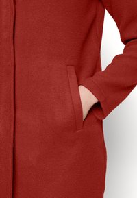 Abrigo de lana rojo con una textura suave, que presenta bolsillos laterales y un diseño simple y recto. Las mangas tienen una ligera tapered.