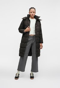 Sieviete, kas uzvilkusi garu melnu pūkaino mēteli pāri baltai īsai džemperi, pelēkām platām biksēm un melniem sporta apaviem, stāv pret vienkāršu fona.