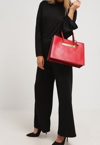 Bolso de piel roja con un diseño estructurado, silueta plana, asas superiores y un acento de hardware en tono dorado. Combinado con un atuendo negro.