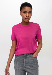 Korte mouw, ronde hals T-shirt van felroze katoen. Model draagt een grijze jeans en staat tegen een witte achtergrond.