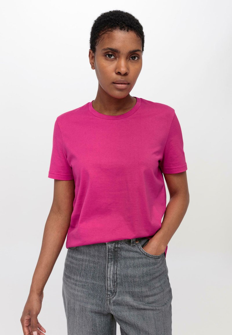Korte mouw, ronde hals T-shirt van felroze katoen. Model draagt een grijze jeans en staat tegen een witte achtergrond.