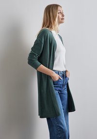 Lång grön kofta i mjukt tyg, med öppen frontdesign, ribbade ärmar och en avslappnad passform, tillsammans med en vit topp och blå jeans.