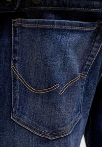 Tylna kieszeń ciemnogranatowych dżinsów z charakterystycznym złotożółtym przeszyciem i zakrzywionymi dekoracyjnymi liniami na materiale.