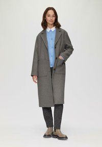 Grijze wollen jas met opstaand kraag, twee voorzakken, laagjes over een lichtblauwe cardigan en zwarte patroonbroek, gecombineerd met enkellaarzen.