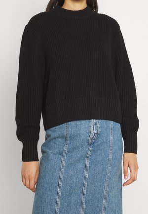 Femme portant un pull en maille noir à manches longues et col montant, associé à une jupe en jean bleue avec des coutures verticales.