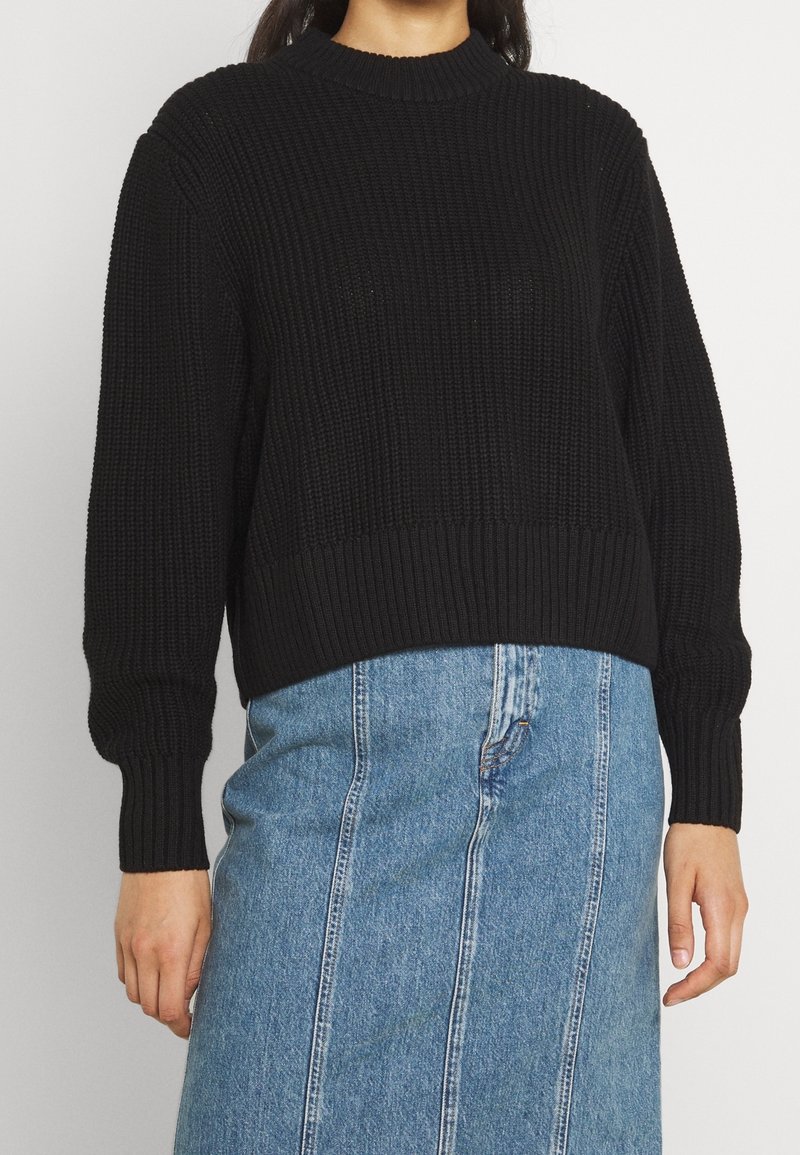 Femme portant un pull en maille noir à manches longues et col montant, associé à une jupe en jean bleue avec des coutures verticales.