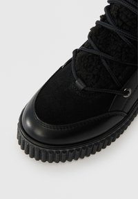 Fekete bokacsizma, velúrból és textúrált anyagból készült felsőrésszel, kerek orral, vastag bordázott talppal és fekete fűzővel, amely fém szemeken keresztül van átfűzve.