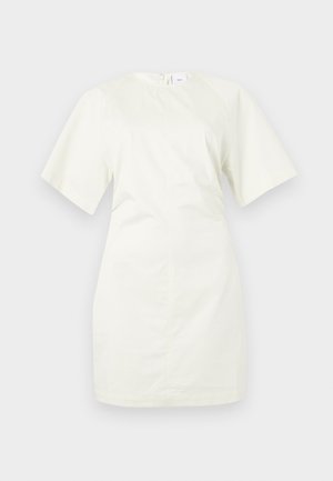 Žena stojaca v krémovom krátkom minináhdeli, čierny dvojitý opasok, strieborný retiazkový náhrdelník a čierne sandále na vysokom podrážke.