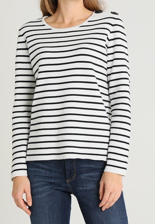 Femme portant un haut à manches longues blanc avec des rayures horizontales noires et des détails de boutons sur les épaules, associé à un jean bleu foncé.