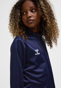 Meisje met krullend haar dat een marineblauw Hummel jas met rits draagt, kijkt naar links tegen een effen achtergrond.