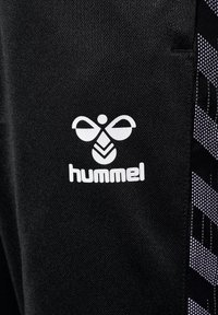 Zwarte stof heeft een opvallend wit "hummel" logo met een gestileerd bijenontwerp. Zijkanten zijn versierd met een geometrisch patroon in paarse tinten.