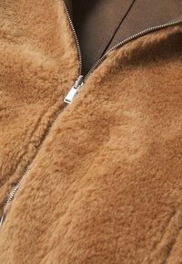 Veste en tissu marron duveteux avec une fermeture éclair argentée partiellement ouverte, dévoilant une doublure intérieure lisse d'un brun plus foncé en dessous.