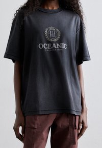 Černé oversized tričko vyrobené z jemné bavlny. Obsahuje grafický potisk s nápisem "OCEANIC EXPLORATION" a dekorativní design koruny. Krátké rukávy.