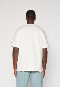 T-shirt en coton blanc avec un col rond, des manches courtes et une coupe décontractée, comportant un petit logo noir sur la manche gauche.