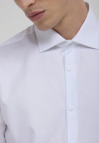 Camicia bianca a maniche lunghe con trama, colletto a punte, dotata di chiusura anteriore con bottoni abbinati, mostrata dalla vita in su.