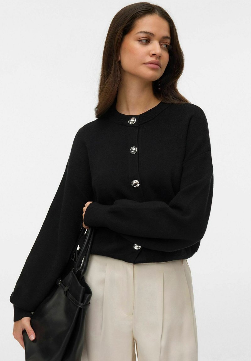 Cárdigan de punto negro con escote redondo, botones plateados y mangas anchas, combinado con pantalones beige de tiro alto y un bolso negro.