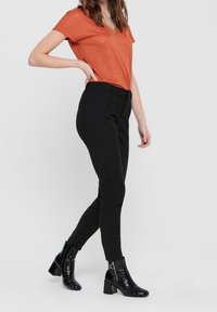 Pomarańczowa koszulka z dekoltem w serek zestawiona z czarnymi zwężającymi się spodniami oraz czarnymi lakierowanymi botkami za kostkę z detalem w formie zamka. Stylizacja podkreśla smukłą sylwetkę.