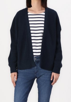 Femme portant un cardigan bleu marine en maille à ouverture devant sur une chemise rayée blanche et bleu marine avec un jean bleu, debout devant un fond blanc.