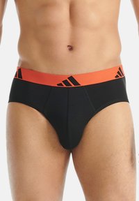 Černé slipy s oranžovým elastickým pasem a designem loga v černé barvě. Tkanina vypadá hladce, má pohodlný střih a minimální švy.