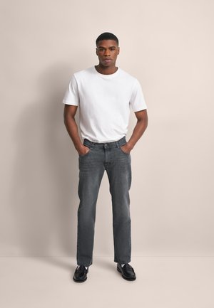 Junger Mann mit Händen in den Taschen, trägt ein weißes T-Shirt, graue Jeans und schwarze Schuhe vor einem einfachen beige Hintergrund.