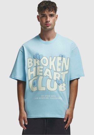 Light blue oversized t-shirt made of cotton featuring bold "BROKEN HEART CLUB" text and blue heart graphics printed on the front.