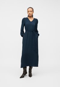 Γυναίκα με πλεξούδες, που φοράει φόρεμα μακρυμάνικο σε ναυτικό μπλε χρώμα με ζώνη στη μέση και μαύρες μπότες με τακούνι, στέκεται μπροστά σε απλό λευκό φόντο.