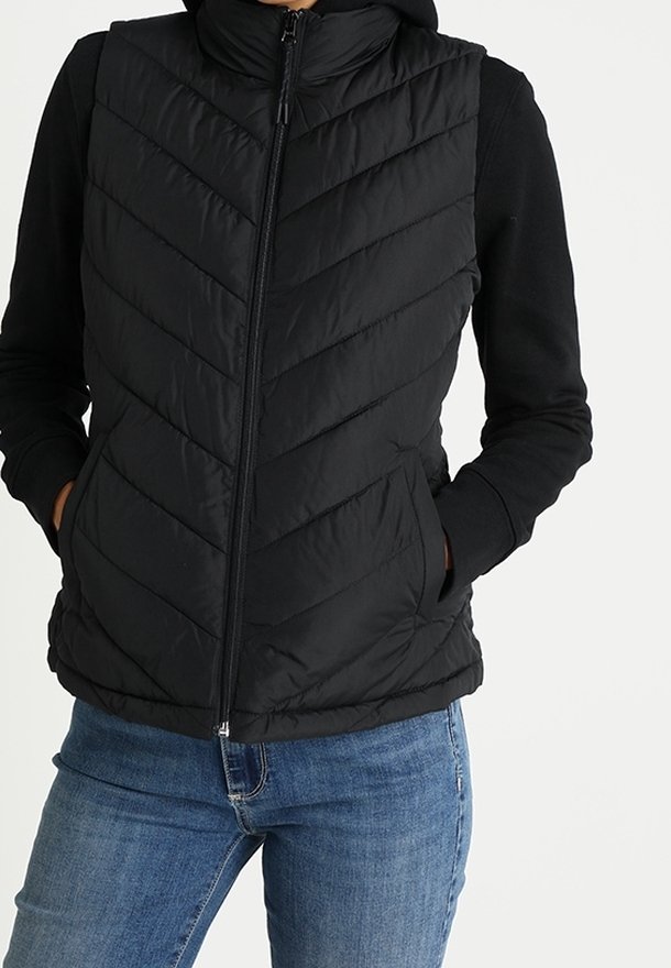 Femme portant un gilet matelassé noir sans manches par-dessus une chemise noire à manches longues, les mains dans les poches du gilet, associée à un jean bleu.