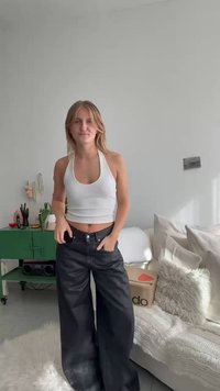 Top curto branco combinado com calças largas pretas, apresentando botões de metal e bolsos. Espaço de vida acolhedor em tons neutros ao fundo.