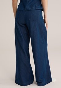 Platas kāju džinsu bikses tumši zilā krāsā, ar aizmugures kabatām un teksturētu apdari. Malas nedaudz ir saplēstas.