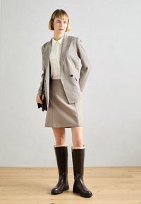 Blazer pied-de-poule avec deux boutons, blouse crème, jupe beige et bottes brunes jusqu'aux genoux avec finition en fausse fourrure et semelles en caoutchouc.