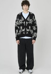Cardigan noir avec un motif floral blanc, texture tricotée, encolure en V et fermeture à boutons, porté par-dessus un t-shirt bleu clair et un pantalon large noir.