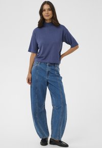 Rövid ujjú, magas nyakú lila felső laza, magas derekú kék farmernadrággal párosítva. Sima textúra és kényelmes szabás. Fényes fekete cipők.