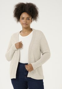 Gilet tricoté beige clair avec une ouverture frontale, des poignets et un ourlet côtelés. Porté sur une chemise blanche, associé à un jeans bleu foncé. Texture lisse.