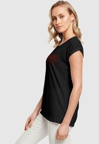 T-shirt noir avec le logo "Stranger Things" en rouge, manches courtes, en coton doux, ras du cou, coupe décontractée, associé à un pantalon clair.